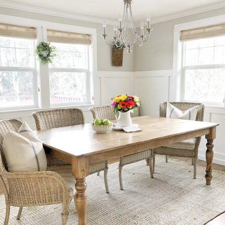 DIY board and batten wainscoting painted white in a dining room with gray paint upper portion of the wall. A wood farmhouse table is centered in the dining room with a pitcher full of colorful florals. Wick chairs surround the table on a light jute rug. 