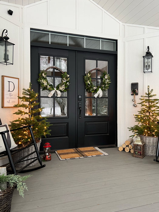 White farmhouse porch with board and batten, shiplap ceiling, wood beams and black lanterns decorated for Christmas. Double black doors decorated with wreaths. Two small Christmas trees on the sides in baskets and with lights. Two black rocking chairs with red and green blankets and pillows. An old wooden sled propped against the wall and a stack of wood beside the sled.