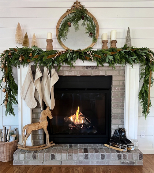 Christmas styled mantel with wooden rocking horse and vintage ice skates on the hearth. Pine and eucalyptus garland draping over the mantel decorated with gold velvet ribbon, gold berries, vintage gold bells and linen stockings. An ornate antique gold mirror is centered on the wall with cream and gold bottle brush trees on one side and wooden candle pillars on the other. A basket of wood sticks sits on the side of the roaring fire.