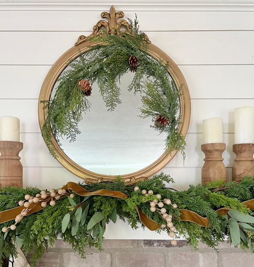 Ornate antique gold mirror with sprig of greenery attached. Wood candle pillars and white candles along the sides. Pine and eucalyptus garlands draped below over the mantel. Vintage gold velvet ribbon and gold berry stems dress the greenery. 