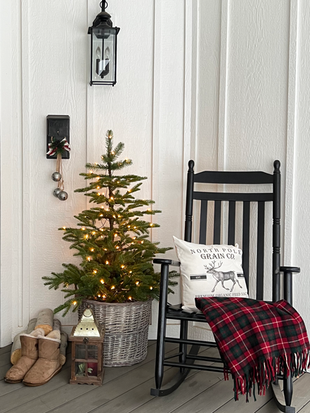 Small lit Christmas tree in a basket with a stack of wood, small lantern, and snow boots next to a black rocking chair, red and green plaid blanket and black and white pillow which reads "north pole grain co."