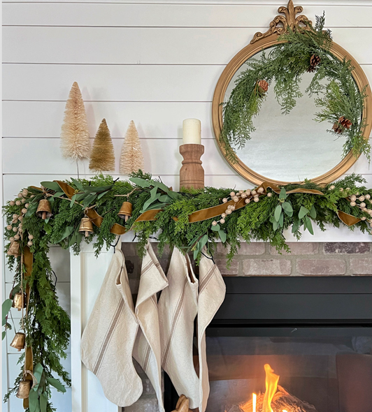 Christmas mantel view with pine and eucalyptus garland and subtle gold heirloom velvet ribbon, bells, and berry stems. An ornate antique gold mirror hangs above the mantel dressed in pine and pinecone wispy garland. Bottle brush trees and wood candle pillars dress the sides. Handmade linen stockings hang to one side in front of a fire.