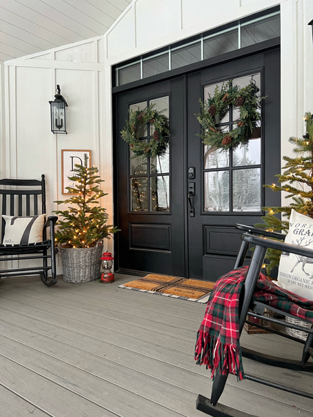 White farmhouse porch with board and batten, shiplap ceiling, wood beams and black lanterns decorated for Christmas. Double black doors decorated with wreaths. Two small Christmas trees on the sides in baskets and with lights. Two black rocking chairs with red and green blankets and pillows. An old wooden sled propped against the wall and a stack of wood beside the sled. 