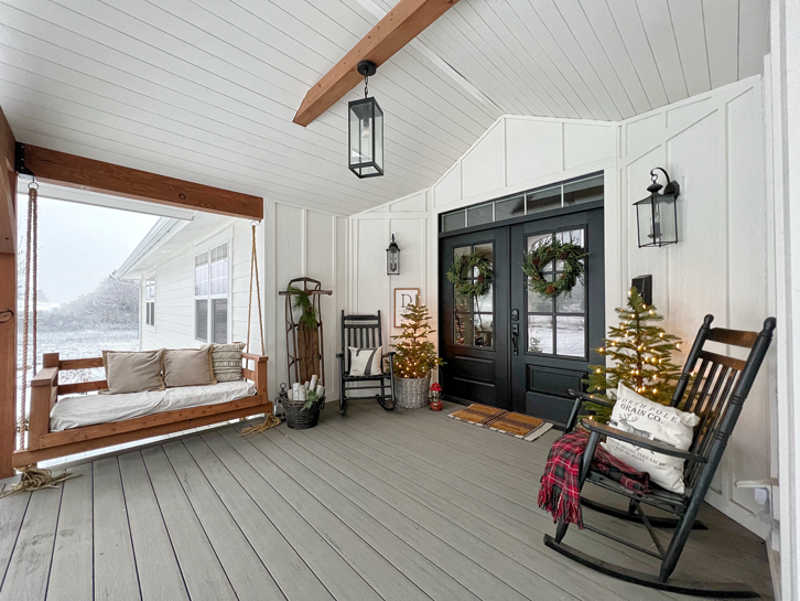 Christmas porch with porch swing, Christmas trees in vintage baskets, a vintage sled, stacks of wood, vintage lanterns and black rocking chairs on a white farmhouse porch with board and batten, wood beams and double black doors with windows
