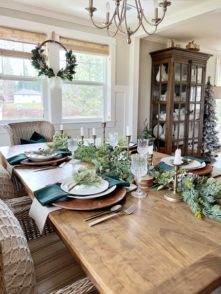 Neutral winter table setting with winter pine and lamb's ear garland as a centerpiece. Centerpiece is filled with brass candlesticks and white candles, white berry stems, and pinecones. Table settings have wooden charger plates, white dishware, and green napkins. Silverware and clear crystal glasses complete the spaces. Table setting sits atop a wooden table. A white shiplap wall and barndoor are in the background.