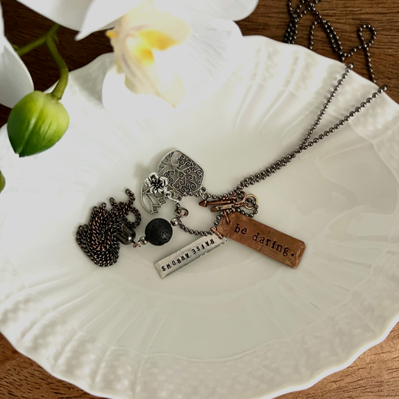 Warmhearted DIY Story Necklaces: Bead ball chain necklace in white bowl with charms. Charms include: two arrows, a pendant reading "raise arrows", tree of life charm, and lotus flower charm.