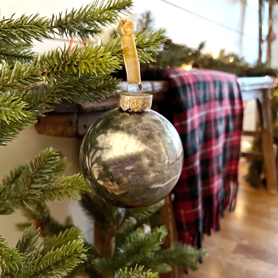 Christmas ornament in twinkling lights on a tree branch with red plaid blanket hanging over a branch in the background. Plastic ornaments that looks like vintage mercury glass in silver and gold. 