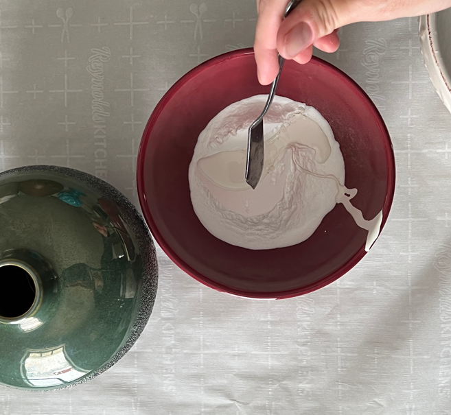 Small piece of pottery on a craft table next to a bowl of baking soda and a spoon full of paint. A hand is mixing the paint and baking soda mixture.