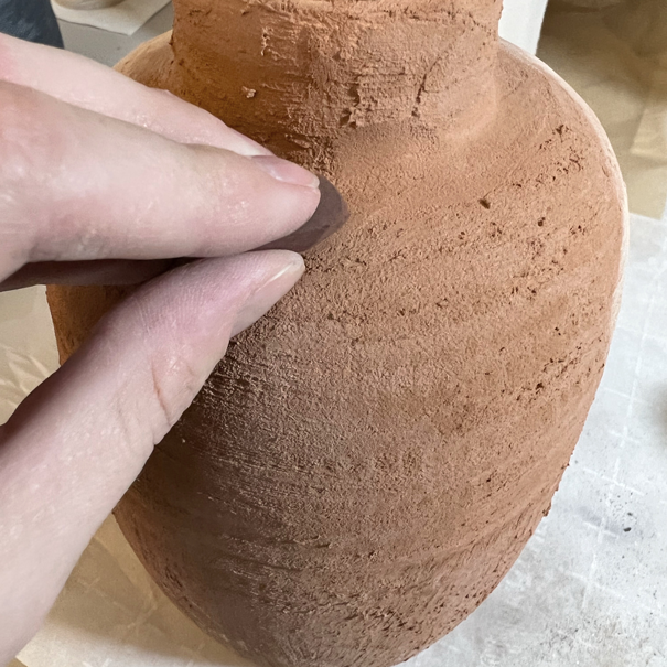 Terracotta colored pottery with rough texture. Hand is adding brown soil-colored chalk.