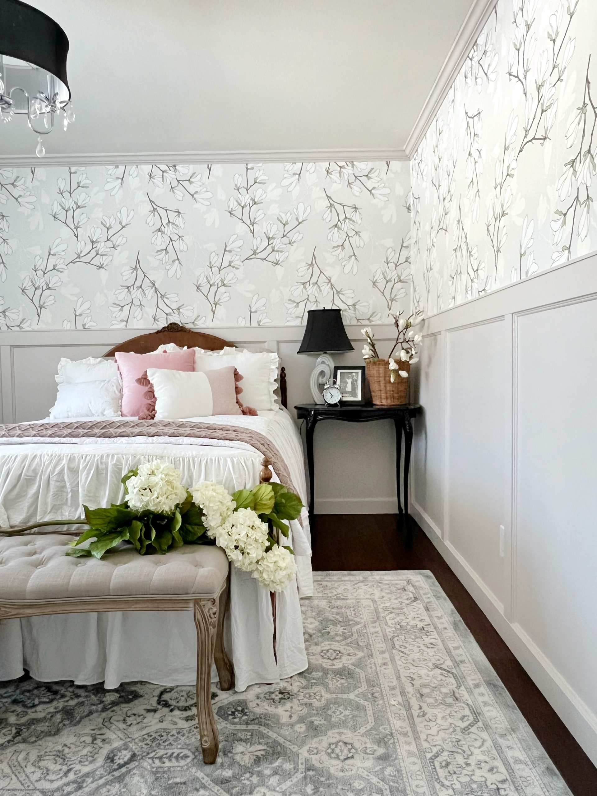 Bedroom with greige board and batten wainscoting and floral wallpaper. A black night stand sits along side the bed with a grey and black lamp, alarm clock and magnolia flowers in a basket. The bed has a ruffled white bedspread.