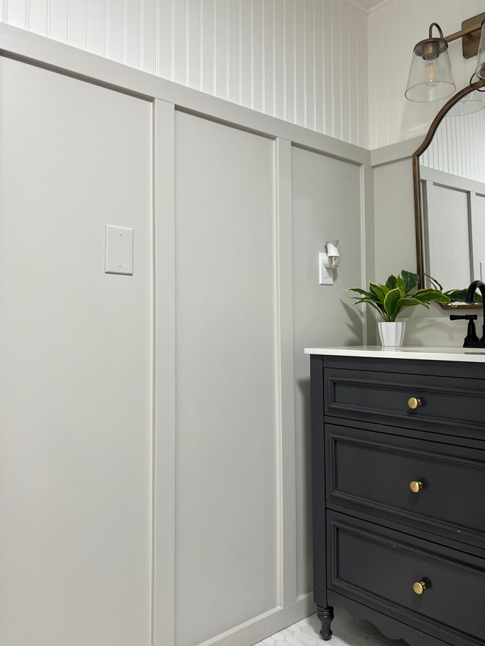 DIY board and batten wainscoting painted light gray in a bathroom with bead board on the top portion of the wall. A black classic vanity and white stone top are featured with a gold mirror and vintage gold lights. 