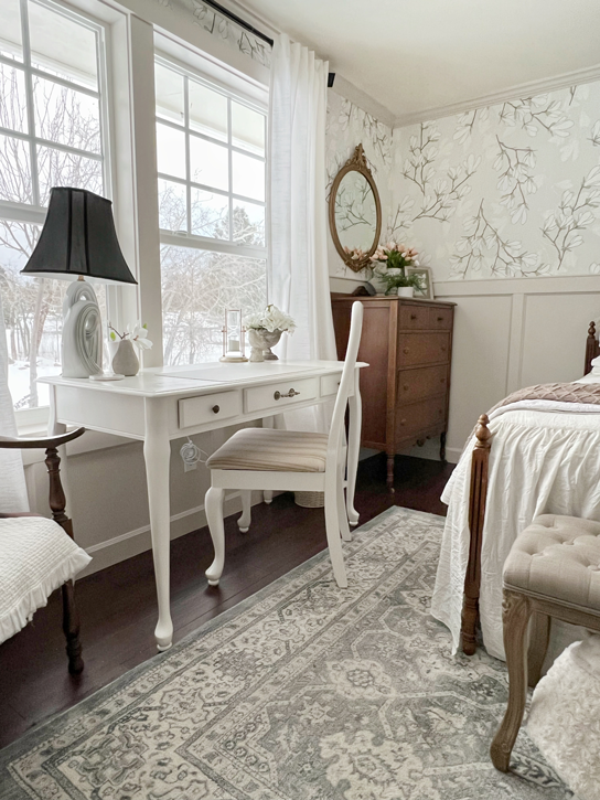 A white desk and chair sit on the left side of the room under big windows of a French country-style bedroom view with board and batten on the lower walls and magnolia floral print wallpaper on the upper portion of the wall.