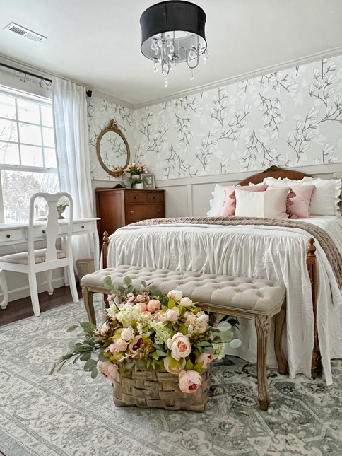 French country-style bedroom view with board and batten on the lower walls and magnolia floral print wallpaper on the upper portion of the wall. Antique arched bed frame with carved embellishments and a matching dresser on the side. Bed covered in flowing white bedding and pink throw pillows. A bench sits at the end of the bed with curvy vintage legs and tufted upholstery. Pink and champagne peonies lay across the bench and a market basket sits under the bench. A white desk and chair sit on the left side of the room under big windows.