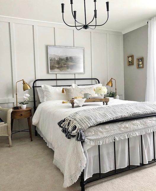 Farmhouse style bedroom with floor to ceiling board and batten. The batten is vertical with one horizontal line 1/3 of the way up the wall. A black iron bed sits in the room with white bedding, and a serving tray styled with a flowers and a breakfast. round wood night stands hold gold lamps on the sides. 