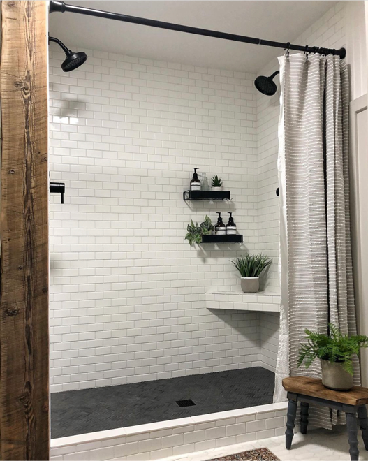 Bathroom shower with white subway tile and matte black fixtures. Matte black herringbone tile covers the shower floor. Black shelving in the shower walls with apothecary shampoo, conditioner, and body wash dispensers. Plants decorate the space both in the shower and on a bench. A rustic wood column lines the shower edge. 