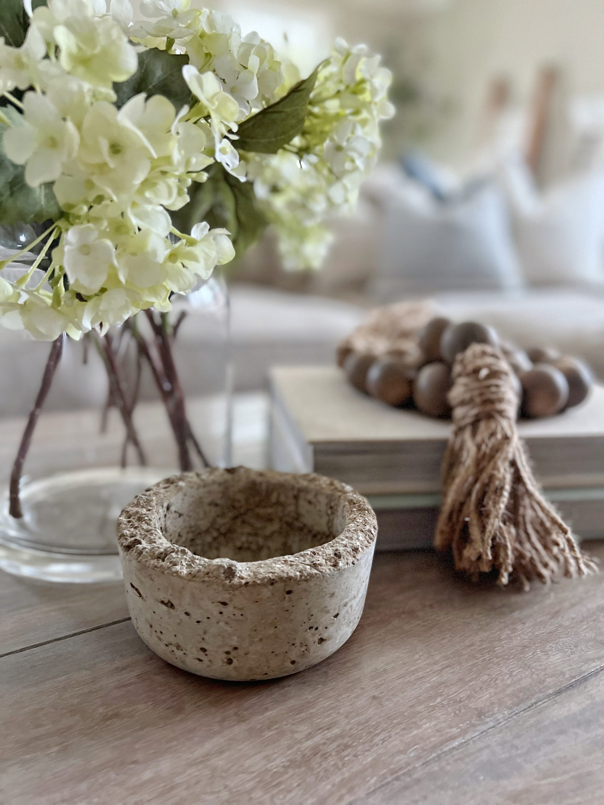 Coffee table centerpiece for easy spring decorating ideas with glass vase of hydrangeas next to a stack of books and wood bead garland draped over the books. A rustic tiny concrete bowl sits in front. 