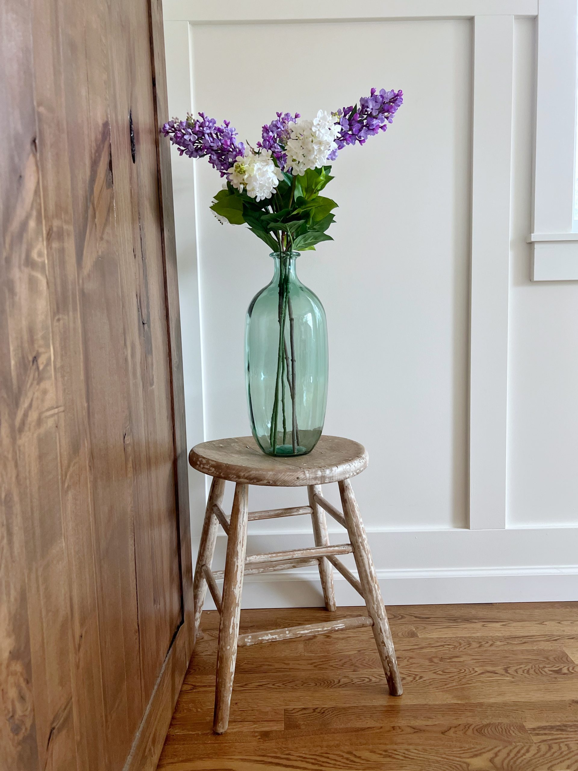 Lilacs in a green glass vase sitting on a round vintage wooden stool.
