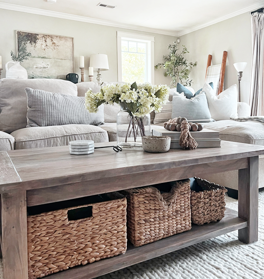 Cozy neutral linen couch scene with subtle blue, beige, and gray throw pillows. Coffee table centerpiece with glass vase of hydrangeas next to a stack of books and wood bead garland draped over the books. A rustic tiny concrete bowl sits in front. Woven baskets sit on the bottom shelf of the coffee table. 
