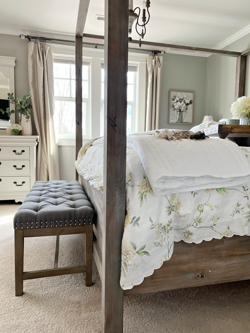 DIY Pottery Barn canopy bed in a king size for a master bedroom. The finish is done a weathered grey and warm brown wood stain. A white duvet with yellow flowers and pale gray-green greenery is on the bed with a white quilted blanket folded at the end and a cashmere alpaca blanket draped over the end. A tufted gray bench sits at the end and a breakfast tray with flowers and a coffee much sit in the middle of the bed. 