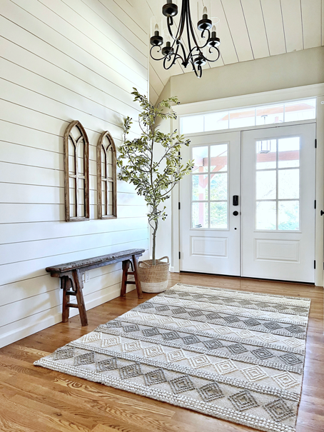 A realistic DIY faux tree is placed in an entryway with double white doors and a transom window over the doors. The ceiling is lined with shiplap and a black chandelier hangs in the center of the room. To the right of the tree a shiplap wall is decorated with a primitive Chinese farmer's bench and arched cathedral windows hang above. On the wood floors is a wool accent rug in grey and white with textured diamond detailing.