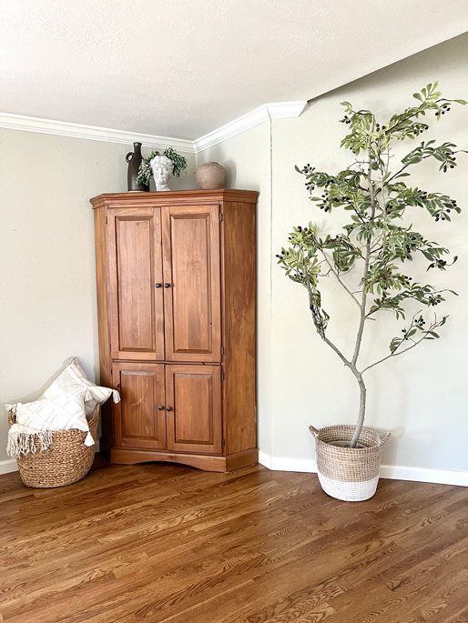 A realistic DIY faux tree is placed in a corner next to a tall wood corner hutch. A basket of throw blankets and pillows sits on the floor in front of the hutch. 