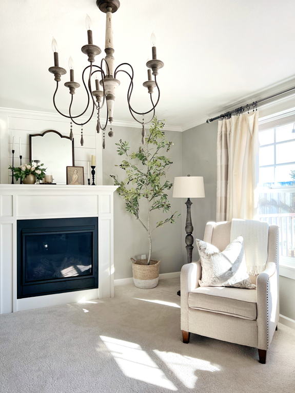 A realistic DIY faux tree is placed in a corner next to a tall floor lamp and a neutral wingback chair with a throw pillow and blanket. On the opposite side is a fireplace painted white against soft gray walls. The mantle is decorated with a vintage arched mirror, black iron candlesticks, a small vintage portrait of a woman in a dress, and a gold vase with pale blush roses and eucalyptus. Centered in the scene and closer in view is a white and wood vintage chandelier. 