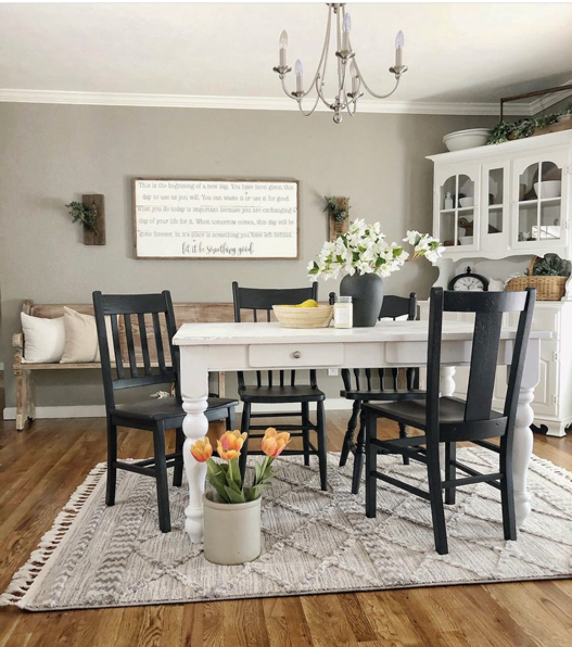 Dining room with white farmhouse table and black chairs. A sign flanks the back wall. On the table is a bouquet of magnolia flowers and a woven bowl of lemons. Sitting on the rug is a bouquet of orange tulips in a crock., Behind the table is a white farmhouse style china hutch.