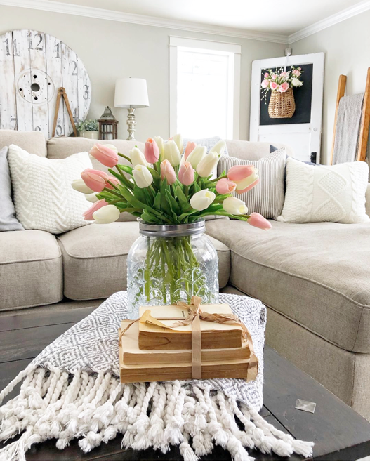 Easy spring decorating ideas. Coffee table in front of a linen colored couch and white throw pillows. In the background is a vintage door with a basket hanging on it filled with pink flowers. In the front is a glass jar full of pink and white tulips and a stack of vintage books.