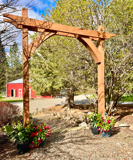 Easy DIY garden arbor with two posts, top beams with curve detail, and curved corbels on a garden path with flowers and a red barn in the background.