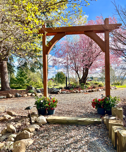 Easy DIY garden arbor with two posts, top beams with curve detail, and curved corbels on a garden path with flowers and a pink flowered cherry tree in the background.