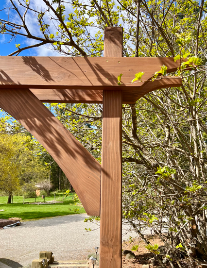 Easy DIY garden arbor up close detail with two posts, top beams with curve detail, and curved corbels on a garden path with flowers and a red barn in the background. 