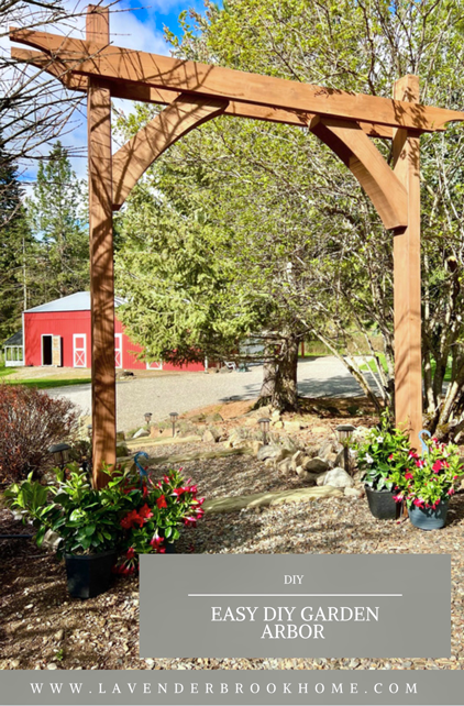 Easy DIY garden arbor with two posts, top beams with curve detail, and curved corbels on a garden path with flowers and a red barn in the background.