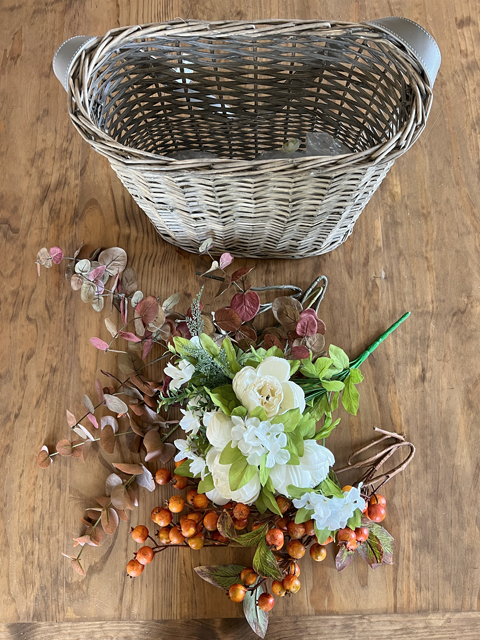 Top view of white peony flowers, orange autumn berry sprigs, and burgundy fall eucalyptus stems, and a wicker market basket. 