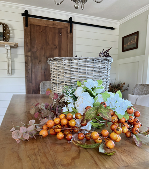 White peony flowers, orange autumn berry sprigs, and burgundy fall eucalyptus stems, and a wicker market basket. 