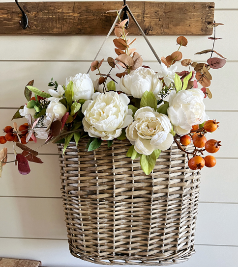 Simple DIY Fall Floral Arrangement made of white peony flowers, orange autumn berry sprigs, and burgundy fall eucalyptus stems hanging in a market basket above a wooden bench. 
