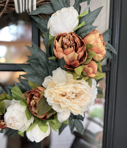 Orange and cream peony wreaths hanging with black and white striped ribbon. The wreaths are made of cream, white, and soft orange peonies arranged in a bed of soft dark green eucalyptus. 