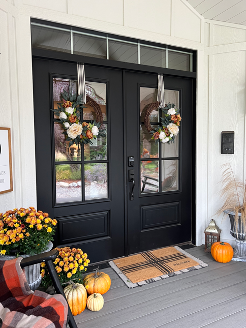 Farmhouse porch with double black doors and orange and cream peony wreaths hanging with black and white striped ribbon. The wreaths are made of cream, white, and soft orange peonies arranged in a bed of soft dark green eucalyptus. 