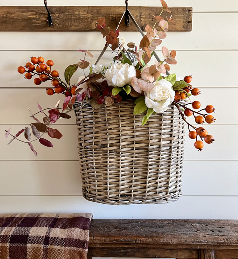 Simple DIY Fall Floral Arrangement made of white peony flowers, orange autumn berry sprigs, and burgundy fall eucalyptus stems hanging in a market basket above a wooden bench.