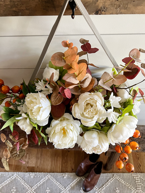 Top view of a Simple DIY Fall Floral Arrangement made of white peony flowers, orange autumn berry sprigs, and burgundy fall eucalyptus stems hanging in a market basket above a wooden bench. 