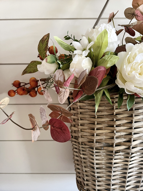 Close up of Simple DIY Fall Floral Arrangement made of white peony flowers, orange autumn berry sprigs, and burgundy fall eucalyptus stems hanging in a market basket. 