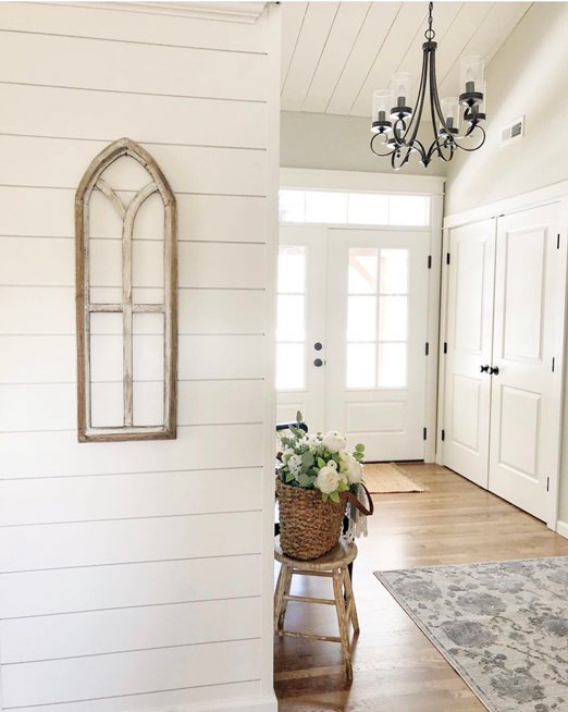 White entryway with double doors. The doors have grided windows and a beautiful transom window above. A basket and bouquet of florals sit atop and small stool with light streaming in.