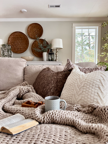 Cozy Hygge-Inspired Winter Living cozy couch scene with a beige chunky woven blanket, knit and faux fur pillows. A cup of tea, a book, and a tic tac toe game rest upon the cozy vignette