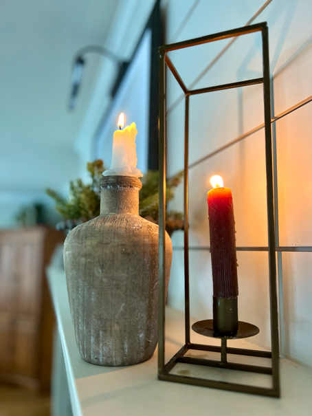 Two lit Cozy Hygge-Inspired Winter Living candle sticks sit upon a fireplace mantel. One within a wooden jug and one within a metal rectangular candle holder. 