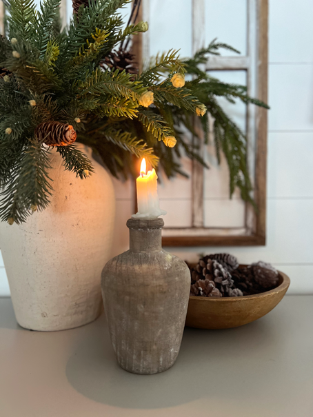 A jug and candle sit in front of a vase of pine branches and a bowl of pinecones. 