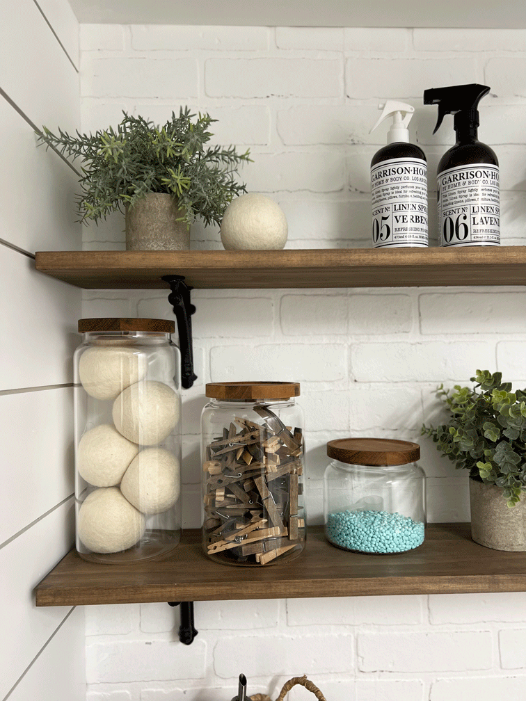 Glass canisters are filled with wool dryer balls, wood clothes pins, and laundry beads. Plants and soap dispensters/ linen sprays in pretty containers also sit atop wood shelving with black iron brackets against a white brick wall. 