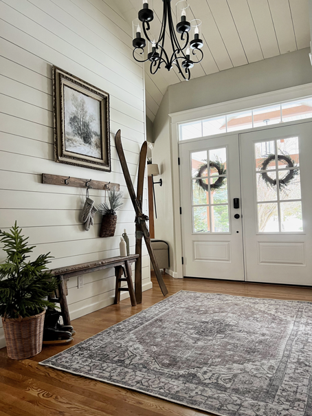 A Cozy Hygge-Inspired Winter Living entryway with double doors is filled with winter decor of skis, a small pine tree, an antique wooden bench, mittens, pine branches, ice skates, and winter art.