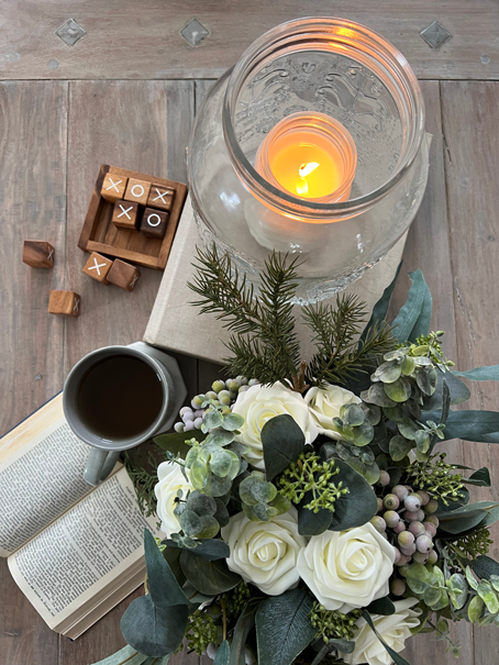 A Cozy Hygge-Inspired Winter Living flatlay view of a cup of tea, an open book, a pine and white rose bouquet, a lit candle glowing from within an embossed jar, and a wooden tic tac toe game. 