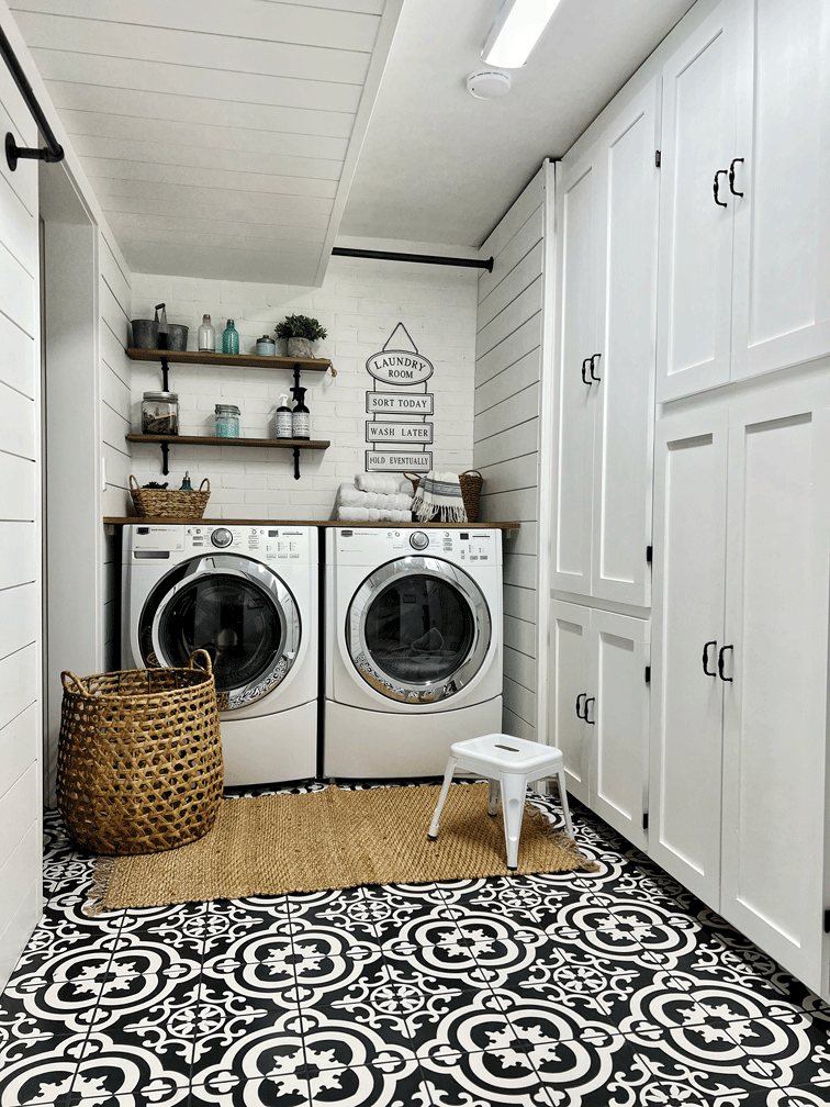 A laundry room with built in storage, bar height folding table, drying racks for hanging or hang drying laundry, dirty laundry metal storage baskets, and decorated with woven baskets, a laundry room sign, a woven rug, and patterned tile to compliment the wood and white board and batten and shiplap walls.