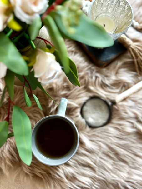 Overview of a cup of hot tea, a magnifying glass, a candle in a votive, and a bouquet or roses and eucalyptus all resting atop a faux fur rug. 