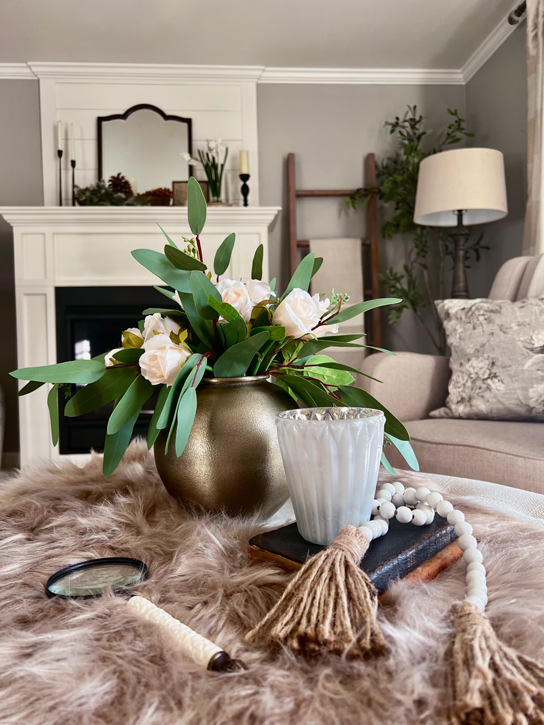 Cozy Hygge-Inspired Winter Living -A view of a cup of hot tea, a magnifying glass, a candle in a votive, and a bouquet or roses and eucalyptus all resting atop a faux fur rug. A side chair sits along side with a mantel and mirror in the background. A blanket ladder, floor lamp and olive tree sit in the corner. 